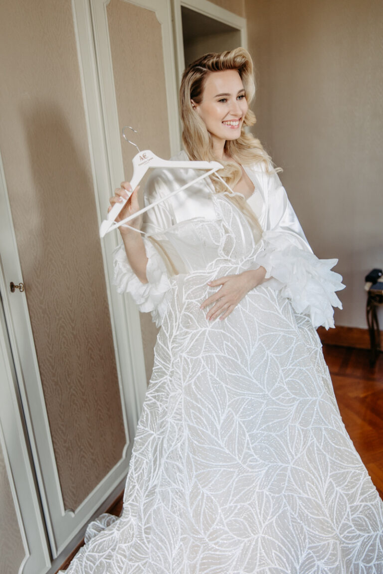 Hochzeitsfotograf Gardasee 10 Strahlende Braut in Satin-Morgenmantel hält ihr Hochzeitskleid auf einem Bügel, voller Vorfreude an ihrem Hochzeitstag am Gardasee.