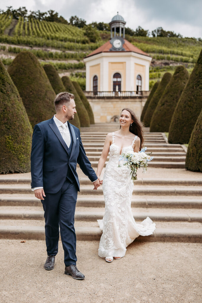 Glückliches Brautpaar spaziert Hand in Hand durch die Weinberge im Schloss Wackerbarth bei Dresdens vor historischer Kulisse.